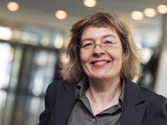 Maria-Sibylla Lotter Eine Person mit welligem blondem Haar trägt einen dunklen Anzug, steht in einem modernen Gebäudeflur mit verschwommenen Lichtern im Hintergrund.