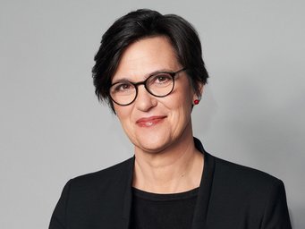 Paula-Irene Villa Braslavsky Oberkörper einer Person in schwarzem Sakko mit einem roten Ohrring vor grauem Hintergrund.