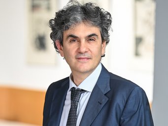 Lucio Baccaro Graumelierter Mann trägt dunkelblauen Anzug mit gepunkteter Krawatte, Hintergrund unscharf mit Kunstwerken an der Wand.