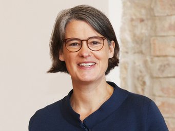 Katrin Kinzelbach Person trägt einen dunkelblauen Blazer, steht vor einer Wand aus hellbraunen Steinziegeln.