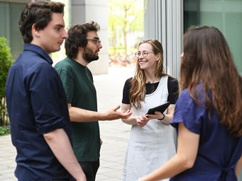 A group of four people conversing near a modern office building, one holding a tablet, with a cityscape background.