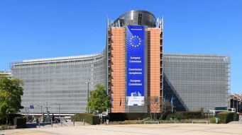 Berlaymont building in Brussels: New legislative initiatives at EU level are usually created in these offices of the European Commission. The building dates back to the late 1960s. The European Commission headquarters in Brussels, showcasing a large banner with the EU emblem and the commission's name in English, French, and German. The building has a modern architectural design with glass and steel elements, surrounded by greenery and a clear blue sky.