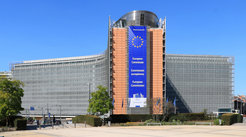 The European Commission headquarters in Brussels, showcasing a large banner with the EU emblem and the commission's name in English, French, and German. The building has a modern architectural design with glass and steel elements, surrounded by greenery and a clear blue sky.
