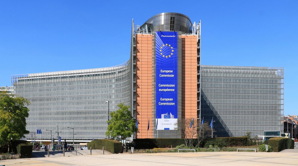 The European Commission headquarters in Brussels, showcasing a large banner with the EU emblem and the commission's name in English, French, and German. The building has a modern architectural design with glass and steel elements, surrounded by greenery and a clear blue sky.