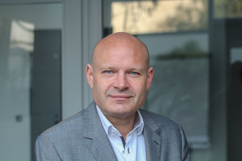 Ein Mann trägt einen grauen Anzug mit einem weißen Hemd und einer dunklen Krawatte, während er vor einer modernen Glasfassade steht.