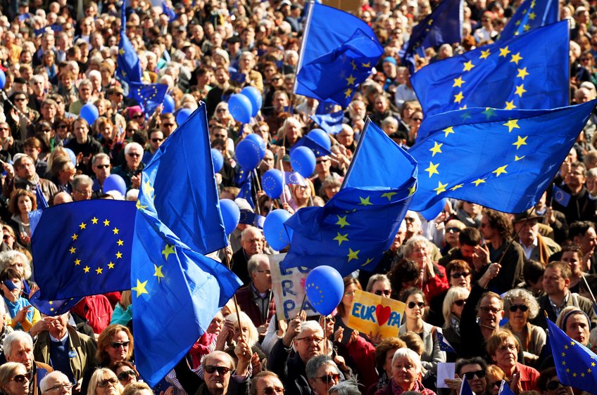 Ist die Europäische Union demokratisch genug? Große Menschenmenge mit EU-Flaggen und blauen Ballons bei einer Veranstaltung im Freien, sonnige Atmosphäre.