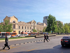 Historisches Gebäude mit klassischer Architektur, umgeben von Bussen und Autos, während Fußgänger bei klarem Himmel die Straße überqueren. Ein Baum und moderne Gebäude im Hintergrund.