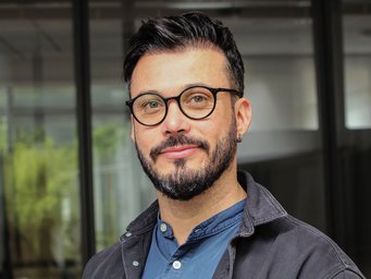 Ab 2019 baute der Wirtschaftssoziologe Felipe González López die erste Partnergruppe des MPIfG in Lateinamerika auf. Die Förderung der Gruppe läuft noch bis Ende 2025. Mann mit Ohrpiercing, schwarzem Haar, in blauem Hemd und dunkler Jacke, vor einem Gebäude mit Glasfenstern.