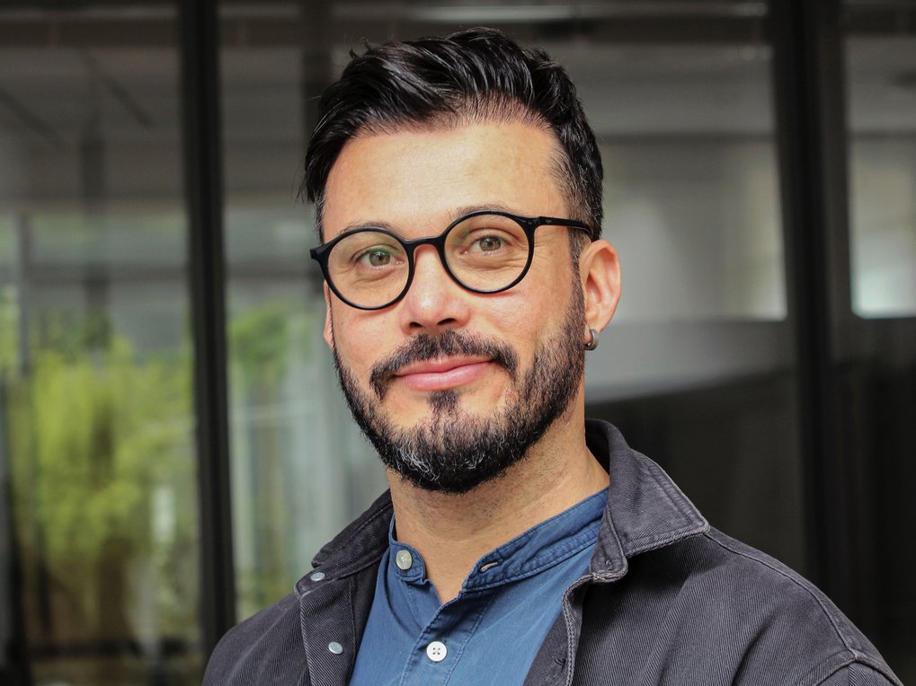 Mann mit Ohrpiercing, schwarzem Haar, in blauem Hemd und dunkler Jacke, vor einem Gebäude mit Glasfenstern.