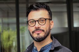 Mann mit Ohrpiercing, schwarzem Haar, in blauem Hemd und dunkler Jacke, vor einem Gebäude mit Glasfenstern.