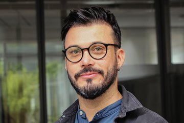 Mann mit Ohrpiercing, schwarzem Haar, in blauem Hemd und dunkler Jacke, vor einem Gebäude mit Glasfenstern.