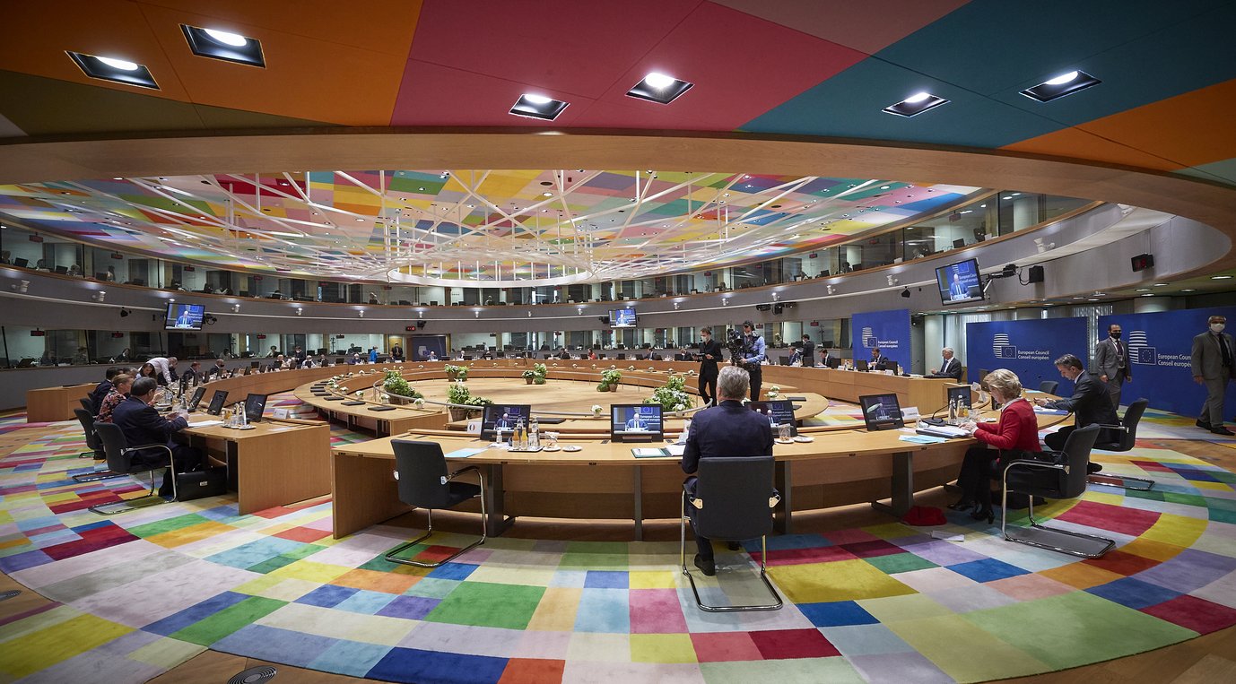 Großer, moderner Konferenzraum mit einem runden Tisch, buntem Teppichboden, mehreren Bildschirmen und Teilnehmern in geschäftlicher Kleidung.