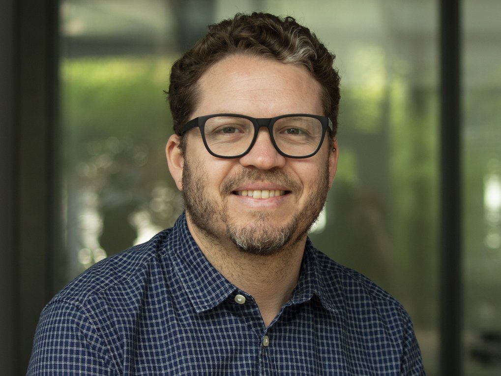 Mann (Aldo Madariaga) mit Brille und in kariertem Hemd vor unscharfem Hintergrund.