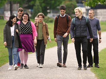 Eine Gruppe von sieben Personen, bestehend aus Männern und Frauen, spaziert in Freizeitkleidung einen breiten Kiesweg entlang, umgeben von grünem Rasen. Das Foto zeigt neue Doktorandinnen und Doktoranden an der IMPRS.