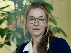 A woman with long, light brown hair, clad in a blue sweater over a white collared shirt, is positioned in front of lush green indoor plants, giving a professional appearance.