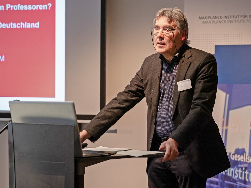 Ein Mann steht auf einem Podium und hält eine Präsentation, während im Hintergrund Folien zu sehen sind.