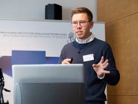 Eine Person hält einen Vortrag auf einer Konferenz, steht vor einem Laptop mit einem Mikrofon und gestikuliert dabei.