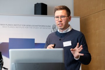 Eine Person hält einen Vortrag auf einer Konferenz, steht vor einem Laptop mit einem Mikrofon und gestikuliert dabei.