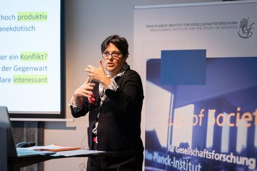 Eine Frau spricht bei einer Präsentation und deutet auf eine Leinwand mit deutschem Text. Sie trägt ein schwarzes Oberteil und hält eine Wasserflasche in der Hand. Hinter ihr befindet sich ein Banner des Max-Planck-Instituts für Gesellschaftsforschung.