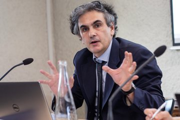 A man in a suit gestures while speaking at a table with a laptop and a water bottle.