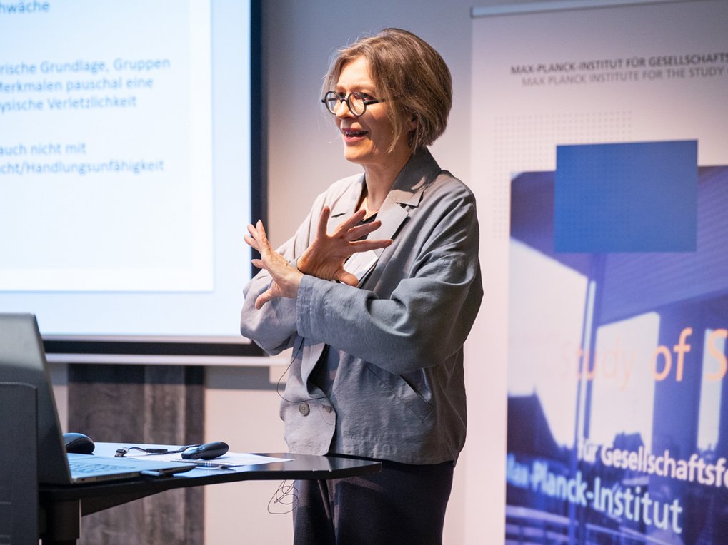 A woman in a gray blazer is giving a presentation, gesturing with her hands, in front of a screen displaying text.
