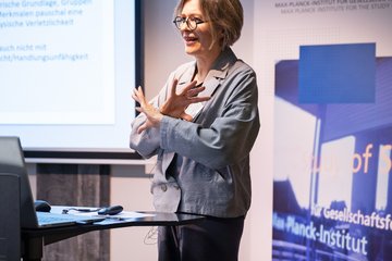 A woman in a gray blazer is giving a presentation, gesturing with her hands, in front of a screen displaying text.