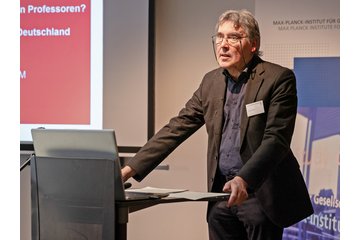 A speaker presenting at a conference or seminar, standing behind a laptop with a slide presentation in the background.