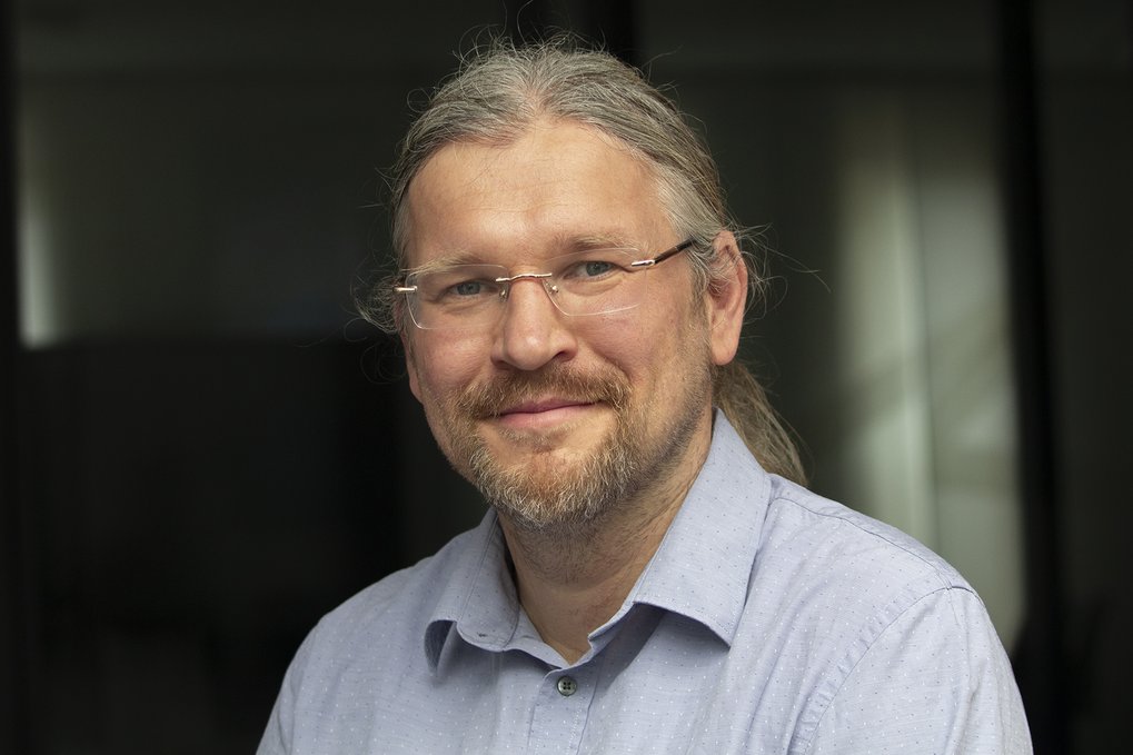 Person in einem hellgrauen Hemd mit Knöpfen, mit langen, nach hinten gebundenen Haaren, sitzt in einem schwach beleuchteten Innenraum mit sanftem Licht, das durch ein Fenster fällt.