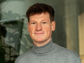 A person is visible in front of a backdrop that consists of plants and blurred architectural details. The person is wearing a grey turtleneck sweater.