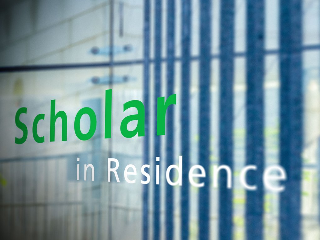 The text "scholar in Residence" is displayed in green and white on a glass surface, reflecting architectural lines and structures in the background.