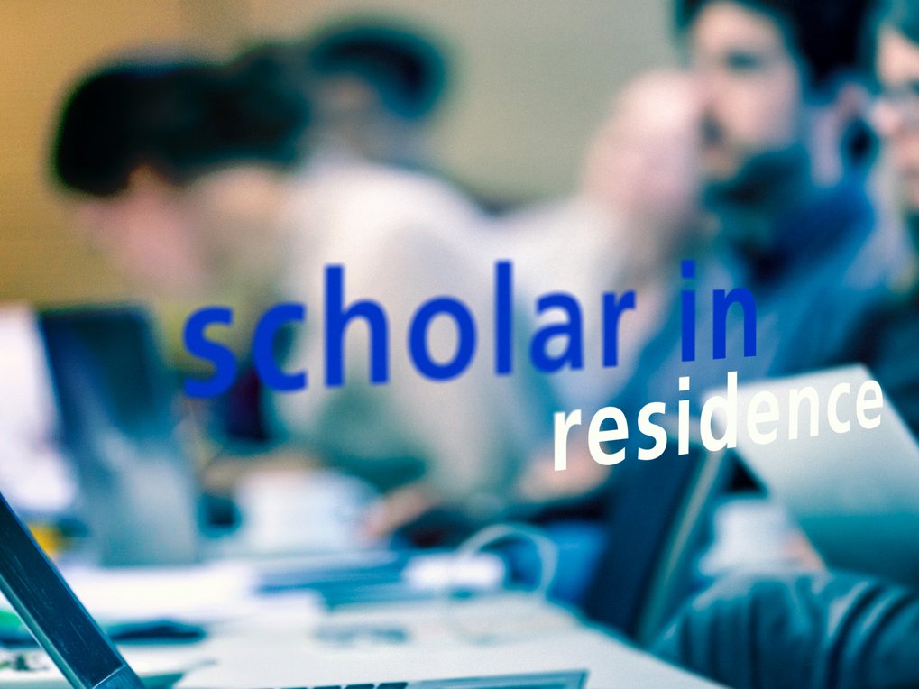 The phrase "scholar in residence" in blue and white is visible on a glass pane, with a blurred background featuring people at desks.