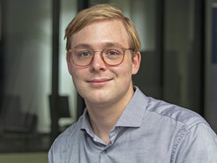 Individual with light blond hair wearing a collared, button-up light blue shirt, positioned indoors, with blurred background elements suggesting a modern office environment.