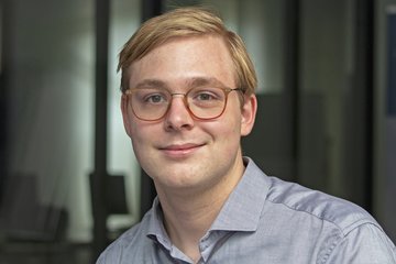 Individual with light blond hair wearing a collared, button-up light blue shirt, positioned indoors, with blurred background elements suggesting a modern office environment.