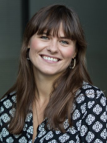 A person with shoulder-length brown hair wears a black blouse adorned with intricate white floral designs, set against a neutral background.