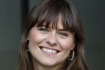 A person with shoulder-length brown hair wears a black blouse adorned with intricate white floral designs, set against a neutral background.