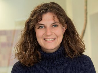 Individual with shoulder-length curly brown hair in a navy blue turtleneck sweater, with small earrings visible, set against a blurred indoor background.