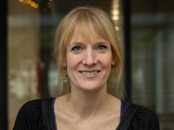 Person with shoulder-length blonde hair, dressed in a black top with a scoop neckline, positioned before a softly blurred background featuring warm ambient lighting and faint reflections.