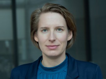 A person named Carola Westermeier, dressed in a blue shirt and a dark blue suit, stands in front of a glass wall that suggests a modern office environment with diffuse lighting conditions.