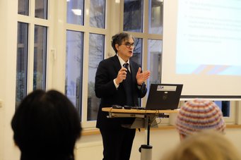 Eine Person steht in einem Raum mit großen Fenstern und hält ein Mikrofon in der Hand. Auf einem Laptop wird eine Präsentationsfolie angezeigt. Im Vordergrund sind einige Teilnehmer zu sehen.