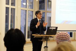 Eine Person steht in einem Raum mit großen Fenstern und hält ein Mikrofon in der Hand. Auf einem Laptop wird eine Präsentationsfolie angezeigt. Im Vordergrund sind einige Teilnehmer zu sehen.