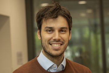 Eine Person in einem braunen Pullover und einem hellblauen Hemd steht vor einem Bürofenster, das eine verschwommene Innenansicht zeigt. Das Foto zeigt Stephan Gruber, der von 2020 bis 2025 Doktorand an der International Max Planck Research School on the Social and Political Constitution of the Economy (IMPRS-SPCE) am MPIfG war.