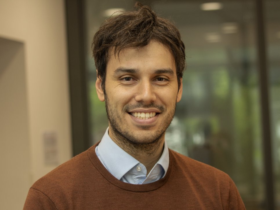 Eine Person in einem braunen Pullover und einem hellblauen Hemd steht vor einem Bürofenster, das eine verschwommene Innenansicht zeigt. Das Foto zeigt Stephan Gruber, der von 2020 bis 2025 Doktorand an der International Max Planck Research School on the Social and Political Constitution of the Economy (IMPRS-SPCE) am MPIfG war.