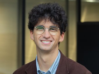 Eine Person mit dunklem, lockigem Haar trägt ein gestreiftes Hemd unter einem braunen Sakko, stehend vor einer Glasfassade.