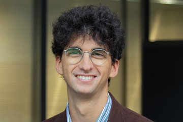 Eine Person mit dunklem, lockigem Haar trägt ein gestreiftes Hemd unter einem braunen Sakko, stehend vor einer Glasfassade.