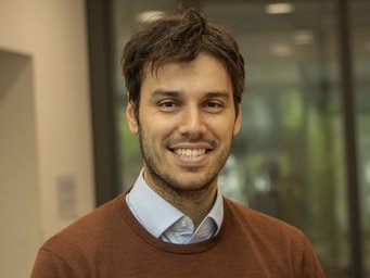 Person wearing a brown sweater over a light blue collared shirt, standing indoors near a glass wall with reflections of greenery outside.