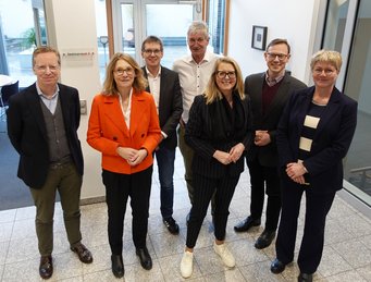 Seven people, four men and three women, stand casually together in the foyer of the MPIfG. They are dressed informally and smile warmly. The scene appears spontaneous and relaxed.