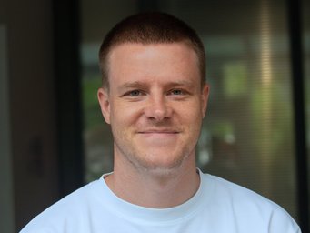 Eine Person mit kurz geschnittenem Haar trägt ein weißes T-Shirt. Der Hintergrund zeigt unscharfe, vertikale Linien und Reflexionen. Das Foto zeigt Dustin Voss, der den Best Paper Prize 2025 der Socio-Economic Review erhalten hat.