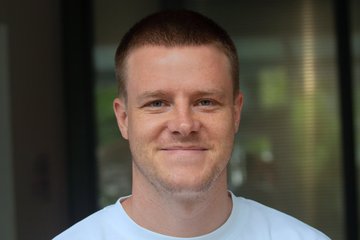 Eine Person mit kurz geschnittenem Haar trägt ein weißes T-Shirt. Der Hintergrund zeigt unscharfe, vertikale Linien und Reflexionen. Das Foto zeigt Dustin Voss, der den Best Paper Prize 2025 der Socio-Economic Review erhalten hat.