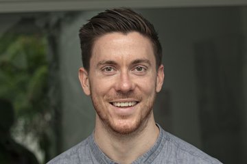 Ein Mann trägt ein graues Hemd mit Knopfleiste; der Hintergrund ist unscharf mit Anzeichen von Pflanzen und Innenraumdetails.