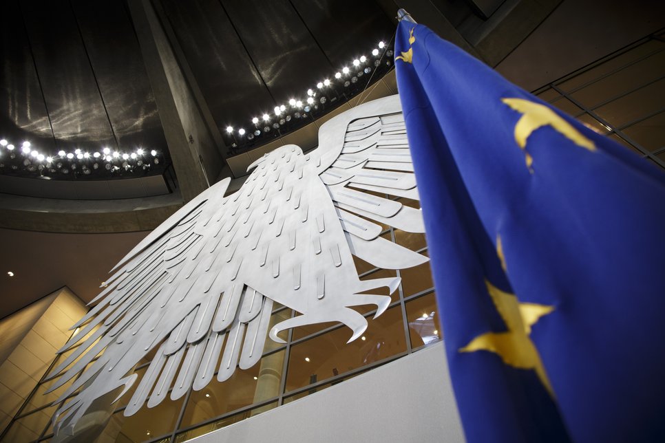 Ein großer Bundesadler hängt an der Wand des Parlamentsgebäudes, daneben weht eine EU-Flagge. Die Decke ist mit Beleuchtung ausgestattet.